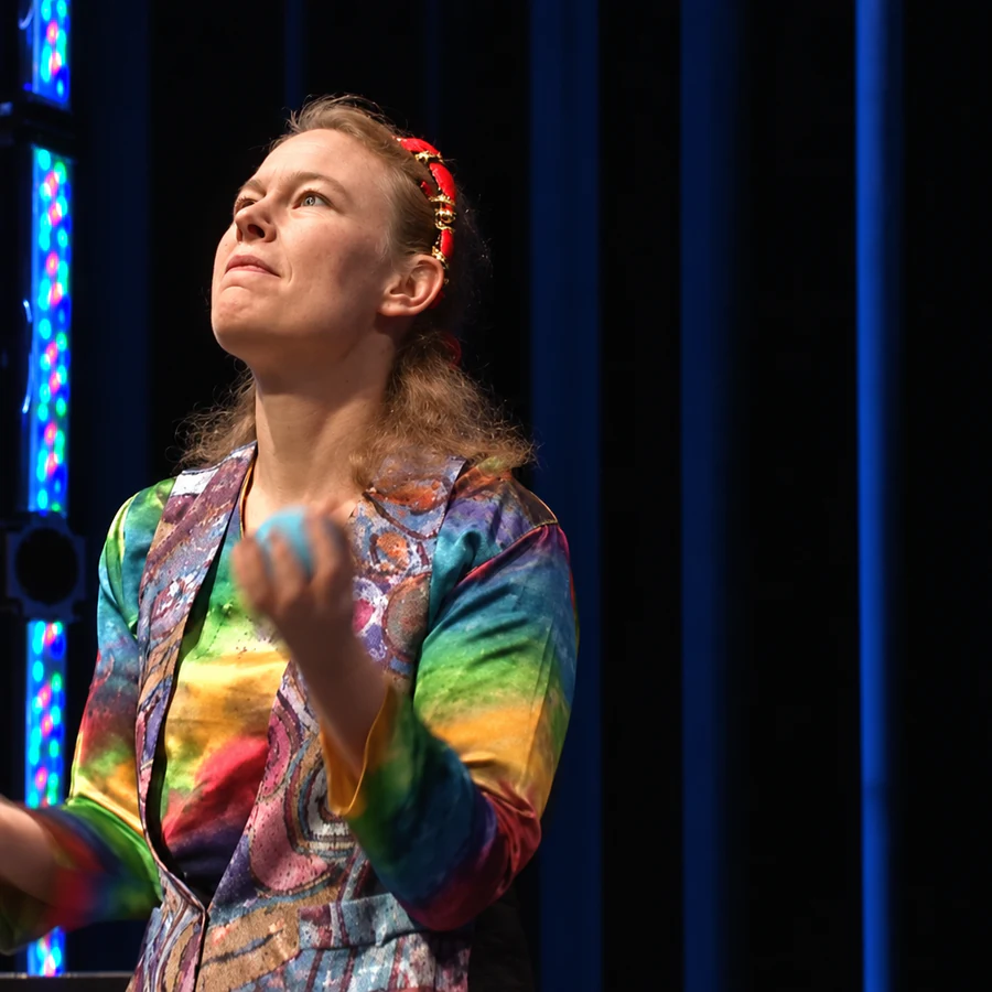 Cindy Marvell juggling in a rainbow tie-dye jacket on the Kennedy Center Millennium Stage